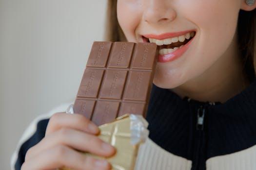 Close-up shot of a smiling woman biting into a chocolate bar indoors.